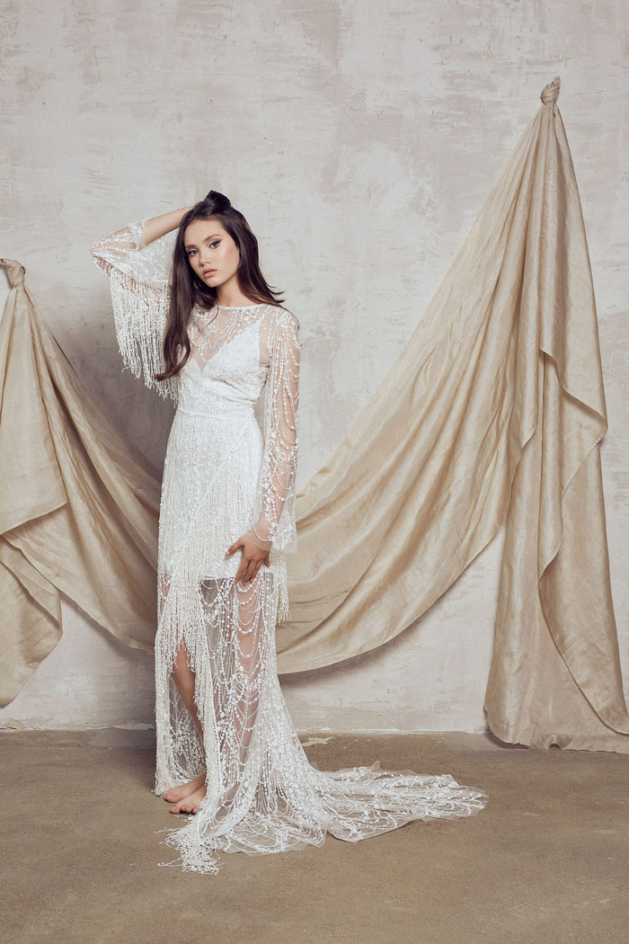 wedding dress designers - wed in florence - BOHEMIAN BEADED LACE EMBELLISHED DRESS WITH LONG BELL SHAPE SLEEVES AND OVERSIZED TASSEL DETAILING. SKIRT CROSSES INTO SPLIT WITH TASSEL HEM.